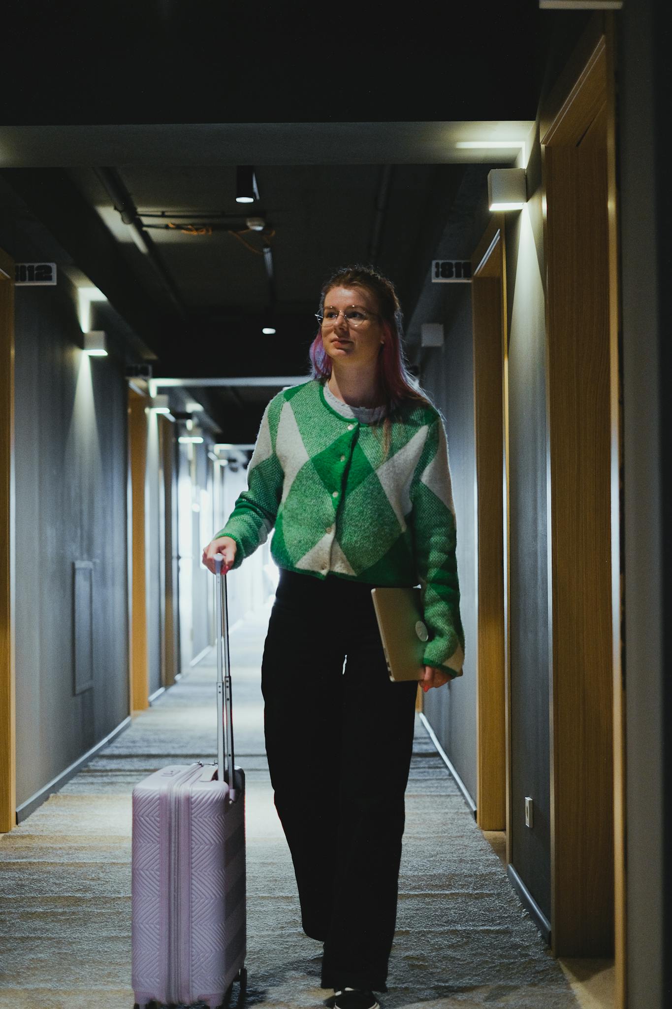 Woman walking through hotel corridor with suitcase, wearing green cardigan.